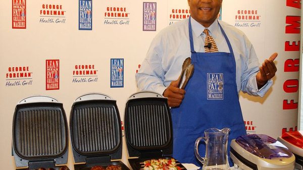 George Foreman presenting his line of grilling appliances, smiling, with various models of the Foreman Grill displayed in front of him.